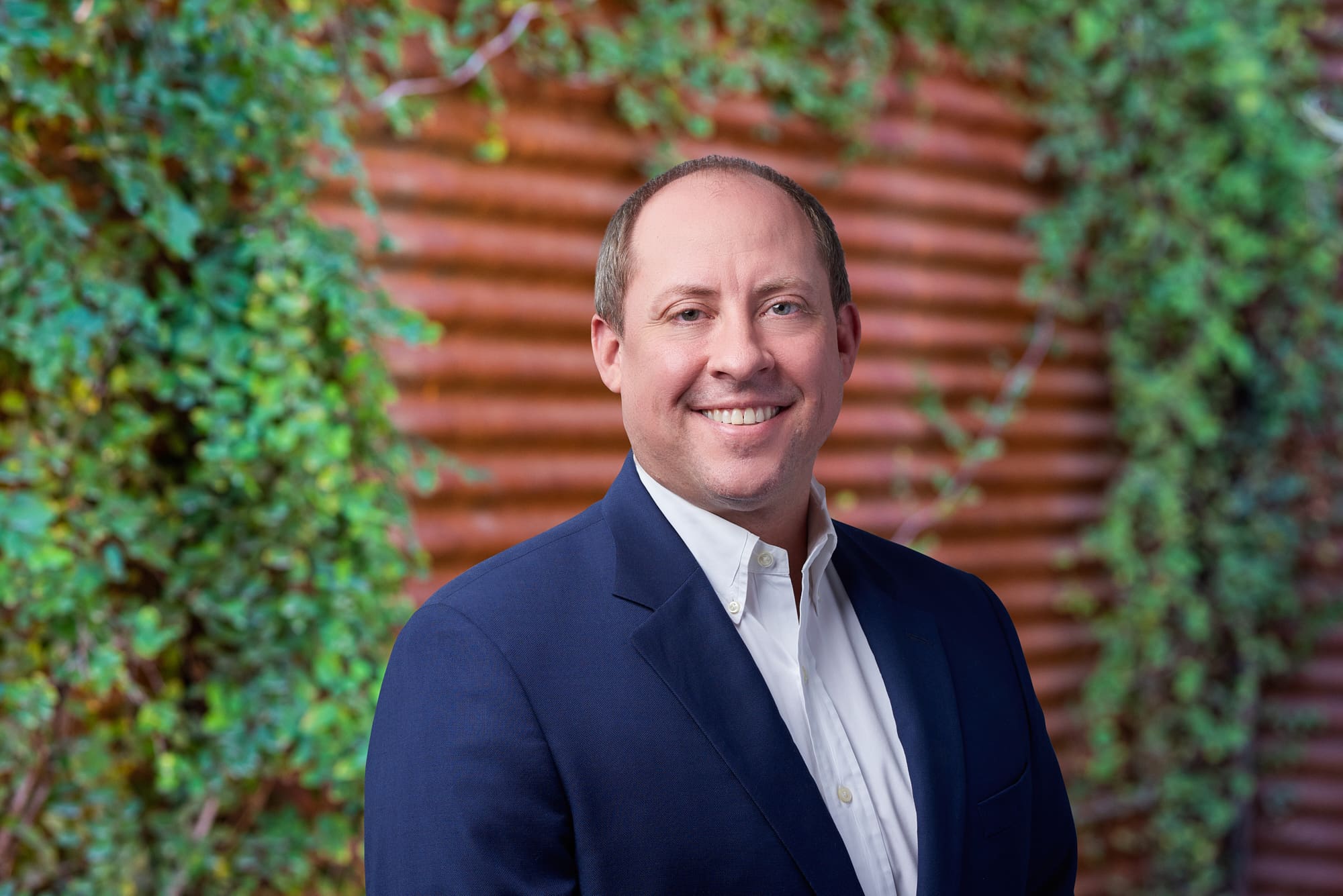 A man named Thomas Jarvis, dressed in a blue suit, smiling in front of an ivy-covered wall.